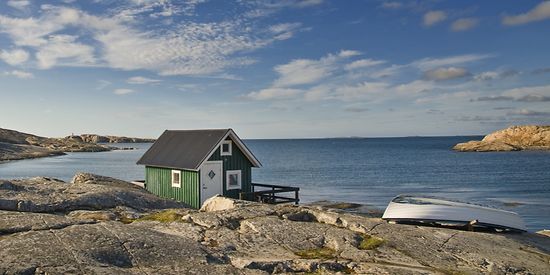 Zusehen ist ein kleines grünes Haus, das sich direkt neben dem Meer befindet. Daneben liegt ein kleines Boot.