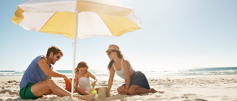 Eine Familie sitzt unter einem Sonnenschirm am Strand und spielen im Sand.